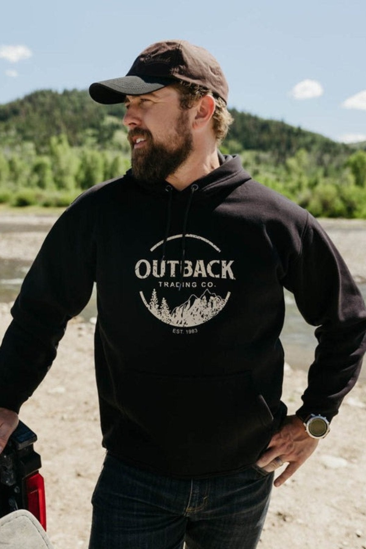 A bearded man in a black cap and Outback Comfy Graphic Hoodie stands by a river with trees and hills in the background, resting one hand on a truck bed.