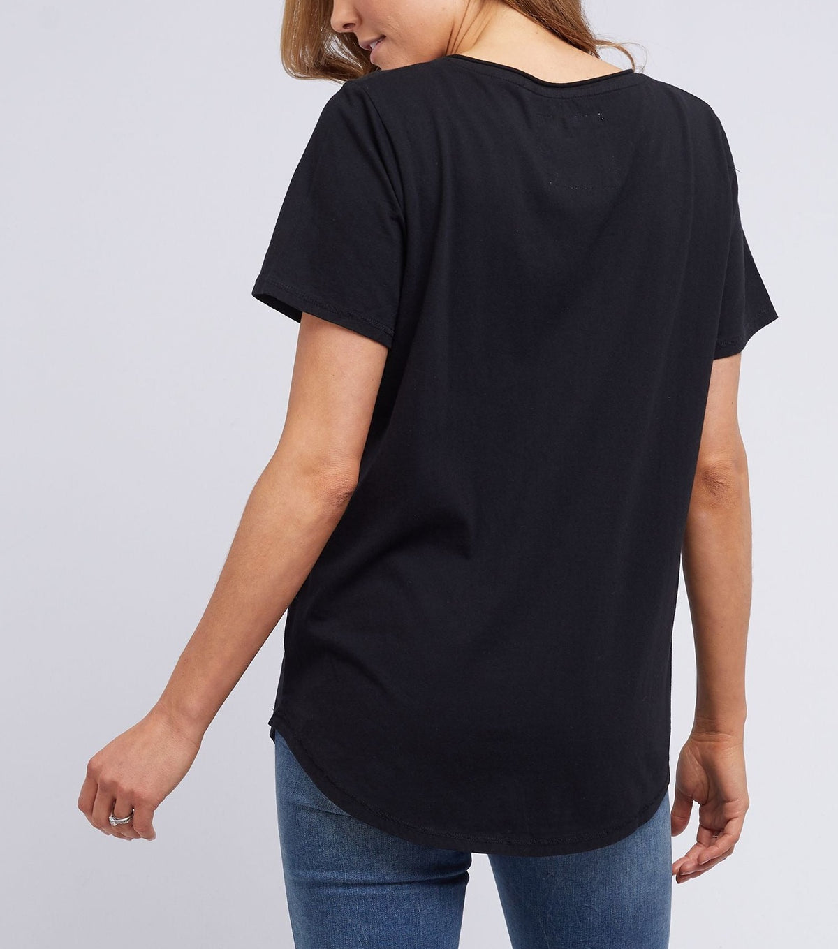 A light brown-haired person wears a black Foxwood Tee, crafted from 100% cotton, paired with blue jeans. From the back, one arm is slightly bent at the elbow against a plain white wall.