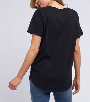 A light brown-haired person wears a black Foxwood Tee, crafted from 100% cotton, paired with blue jeans. From the back, one arm is slightly bent at the elbow against a plain white wall.