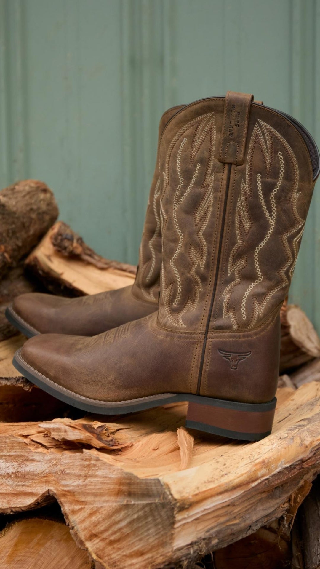 The Pure Western Tolar Boot, featuring brown grained leather and decorative stitching, sits atop split firewood against a greenish wooden wall.