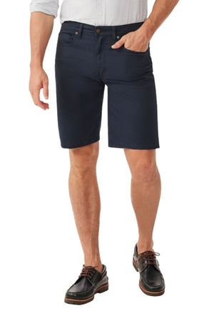 A man stands with one hand in his pocket against a white background, wearing a light long-sleeve shirt tucked into RM Williams Nicholson Shorts and black boat shoes.