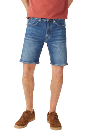Wearing a rust t-shirt, RM Williams Nicholson Short in blue denim, and brown suede sneakers, a person stands with hands out of view; only their lower half is shown against a plain background.