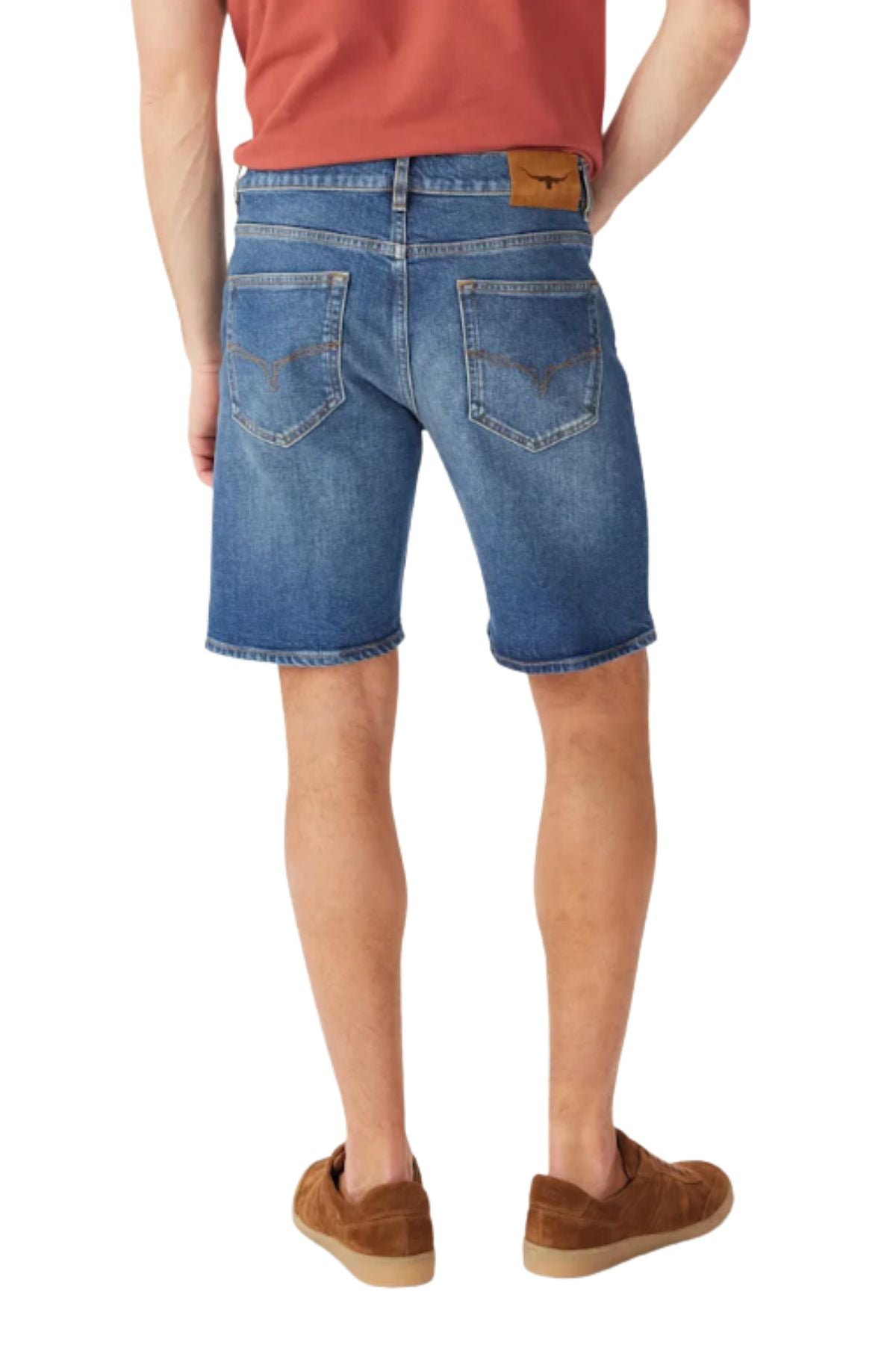 A person stands with their back to the camera against a white background, wearing RM Williams Nicholson Shorts in blue Australian Good Earth Cotton, paired with a red shirt and brown shoes.