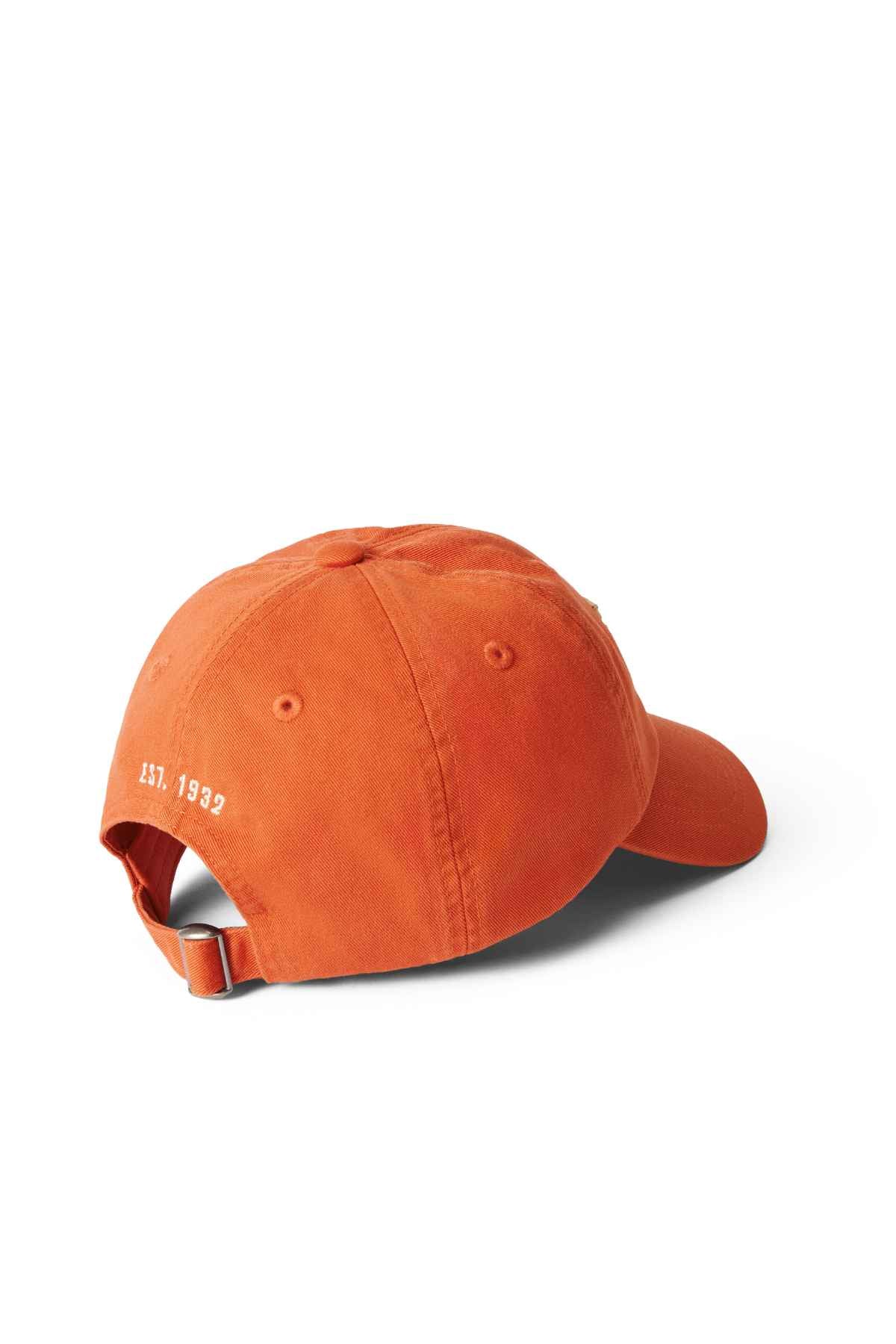 The RM Williams Steers Head Cap, an orange cotton twill accessory, features an adjustable strap with a metal buckle. Elegantly embroidered with "EST. 1938" in white near the strap, it enhances its timeless appeal. The cap is set on a white background and viewed from the back at a slight angle.