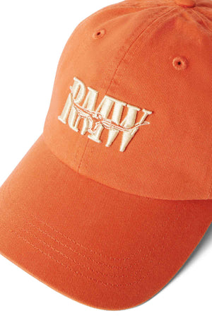 The RM Williams Steers Head Cap is a classic orange cotton twill accessory with a curved brim, featuring cream-colored embroidered lettering and a barbed wire design on the front.