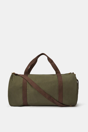The RM Williams Heritage Bag is a green cylindrical duffel made from cotton canvas with brown handles and a matching shoulder strap, featuring a subtle heritage logo print, displayed on a plain white background.