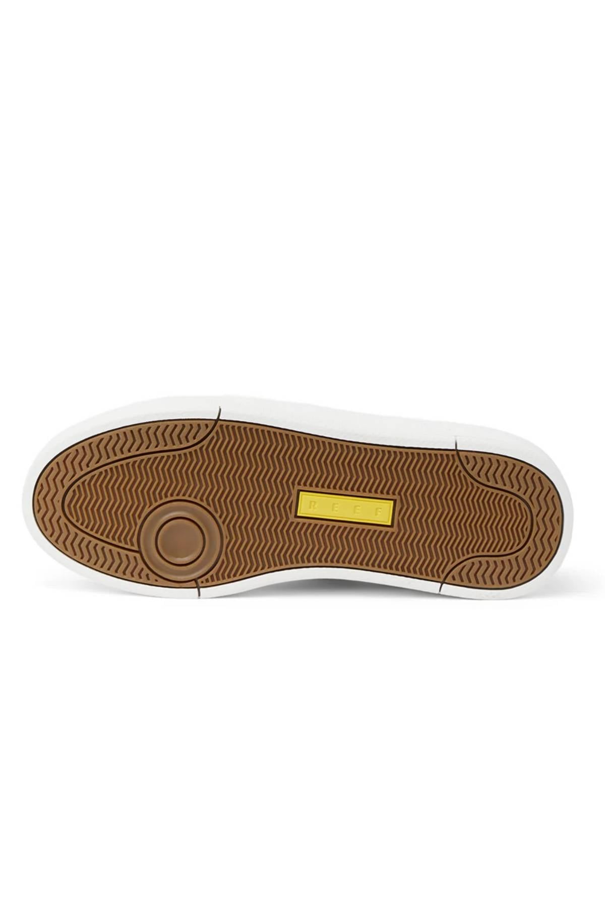 The image shows the bottom view of the Reef Bom Dia Shoe, featuring a brown textured rubber sole with a circular design at the heel, a yellow rectangular label at midfoot, white outer edge, and all-day comfort from its cushioned footbed.