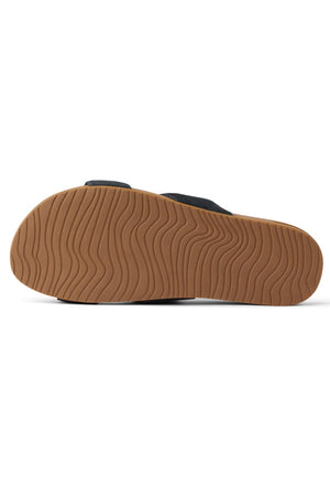 The image shows the bottom view of the REEF Cushion Vista Stevie sandal, featuring a textured, wavy-patterned brown rubber sole and partial black suede upper with built-in arch support for comfort.