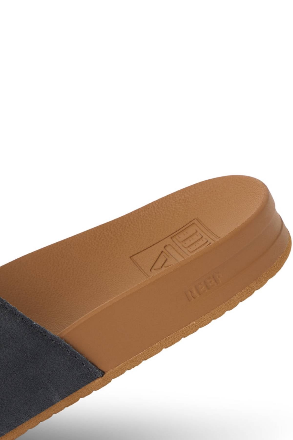 Close-up of the REEF Cushion Vista Stevie sandal featuring brown suede upper, arch support, textured sole, and "REEF" embossed on the side. The upper is dark suede or fabric.