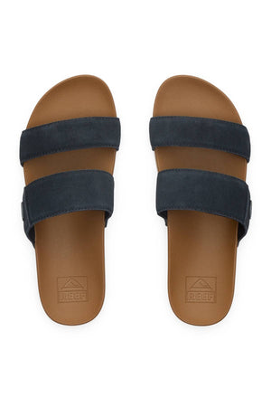 The REEF Cushion Vista Stevie are brown slide sandals featuring cushion midsoles, two wide dark navy blue straps, textured footbeds for comfort, and the "REEF" logo embossed near the heel.