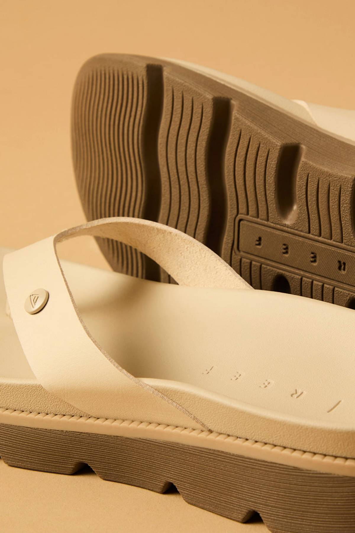 Close-up of two beige REEF Vista Luxe Vivianahh sandals with thick, textured brown soles and wide straps. One sandal lies flat and the other is propped up, showcasing the patterned sole, cushioned footbed, and indented branding on beige.