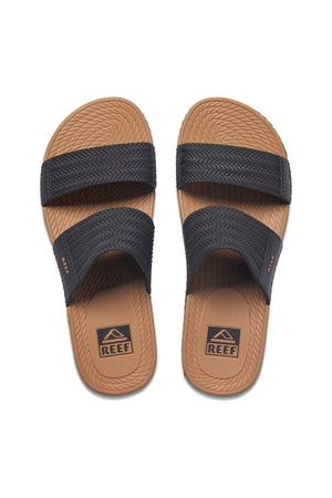 The REEF Water Vista Slide features tan, waterproof sandals with textured soles, two black straps across each foot, and visible "REEF" logos on the footbeds and strap sides. The dual-density EVA footbed provides comfort, while a crisscross pattern adds style.