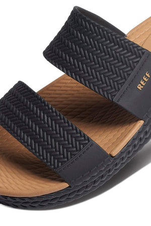 Close-up of a REEF Water Vista Slide sandal in black, showcasing two woven textured straps. It features a brown dual-density EVA footbed, the "REEF" brand on the side of the lower strap, and a textured sole for grip. The sandal is also waterproof.