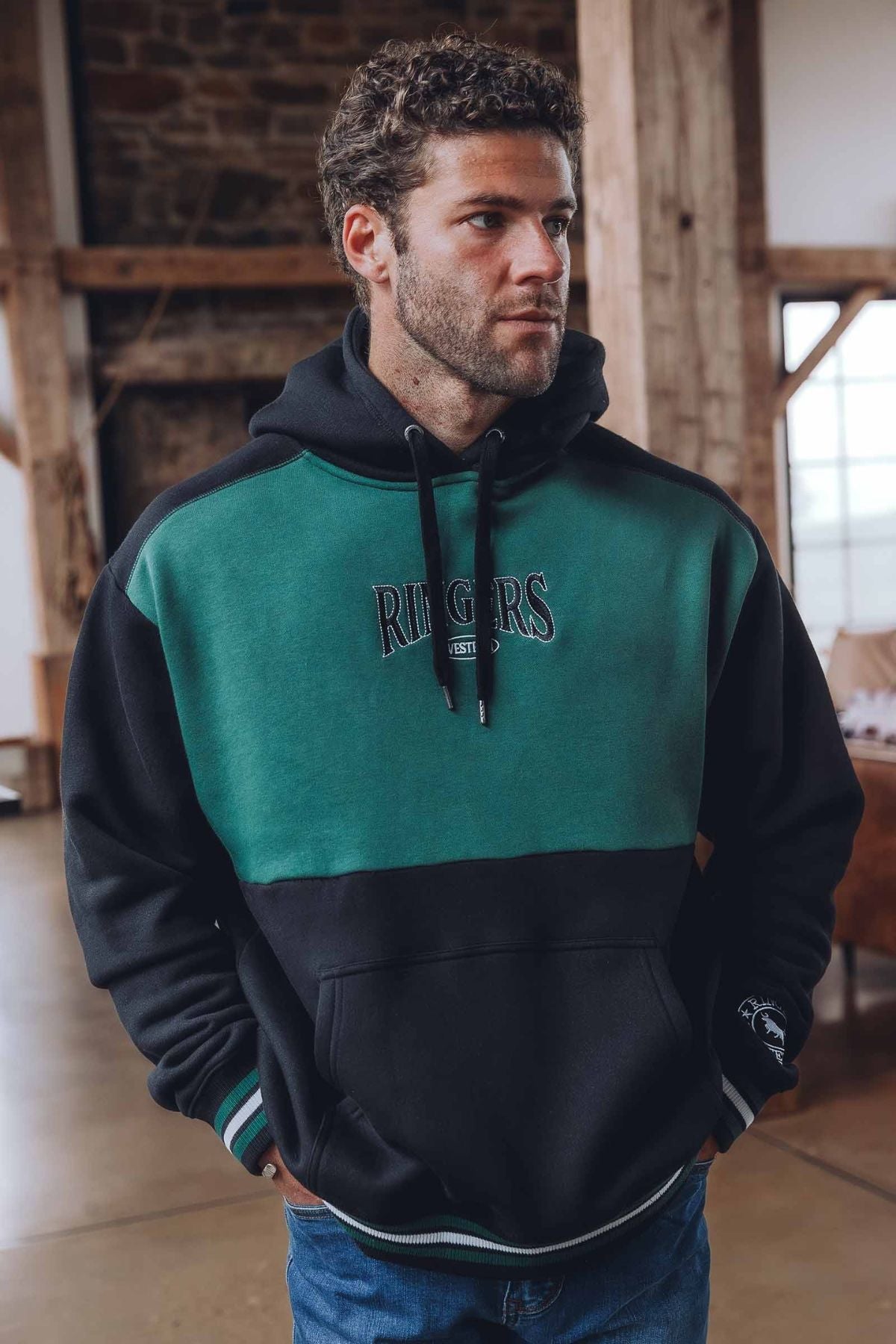 A man with curly hair wears the Ringers Western Barrow Creek Pullover Hoodie in green and black, standing indoors with wooden beams and a stone wall in the background, showcasing the hoodie’s relaxed, casual style.