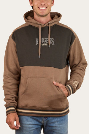 A man wearing the Ringers Western Barrow Creek Pullover Hoodie, featuring a two-tone brown and dark olive design with "RANGERS WESTERN" across the chest, striped cuffs and hem, and blue jeans stands smiling with one hand in the pocket.