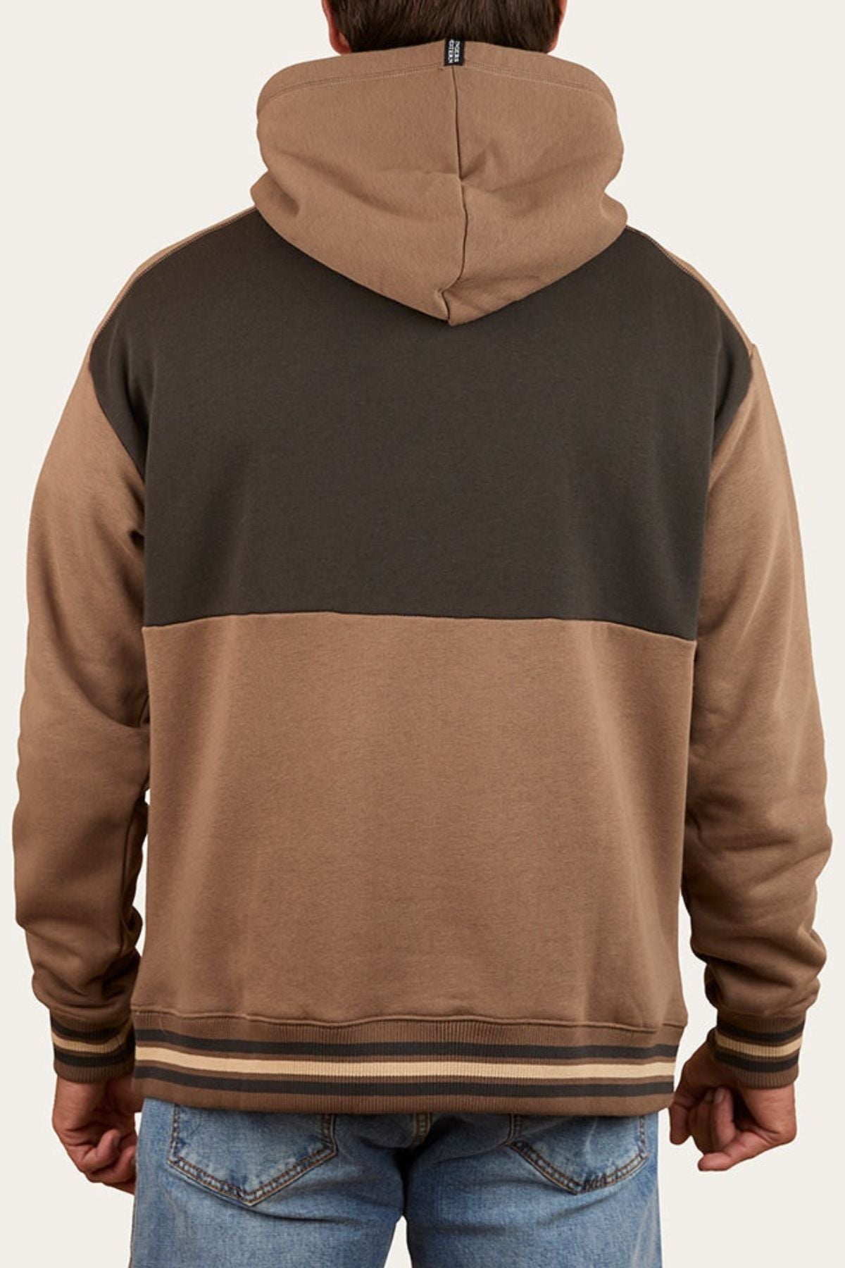 A person in the Ringers Western Barrow Creek Pullover Hoodie, featuring a dark brown upper, light brown lower section, striped cuffs, and an oversized fit, stands with their back to the camera wearing light blue jeans.