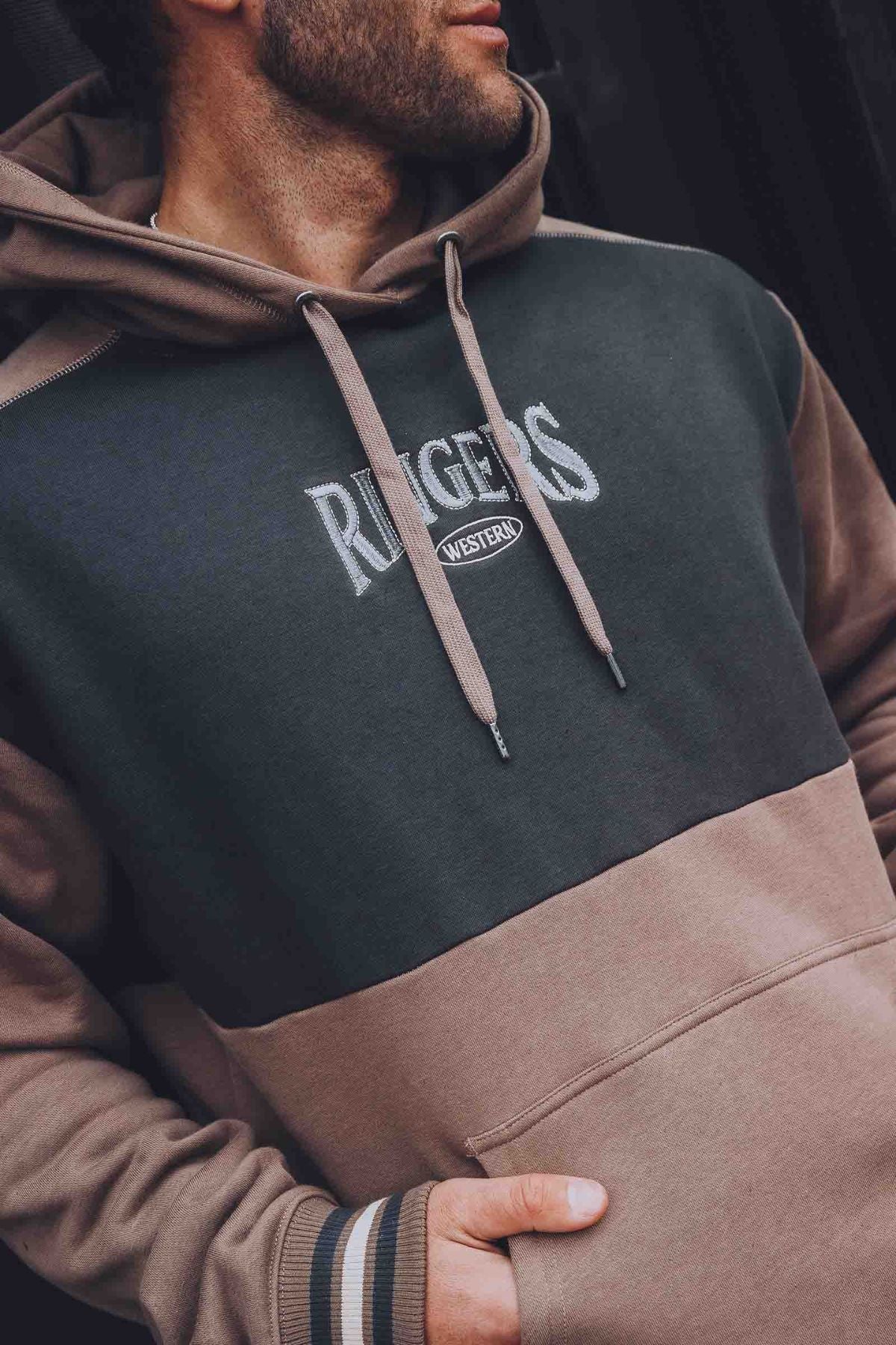 A man wears the Ringers Western Barrow Creek Pullover Hoodie, featuring "RANGERS WESTERN" embroidery on the front in an oversized fit, with one hand in the pocket and glancing slightly to the side.