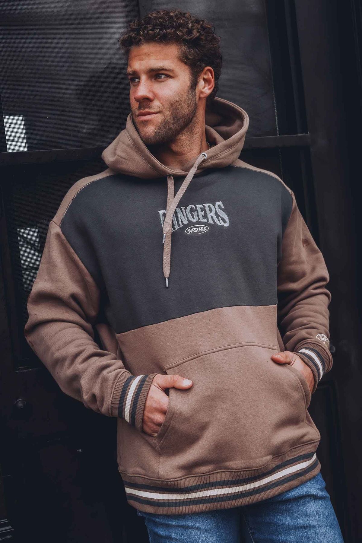 A man with curly hair stands with hands in pockets, looking to the side, wearing an oversized Ringers Western Barrow Creek Pullover Hoodie against a dark background.