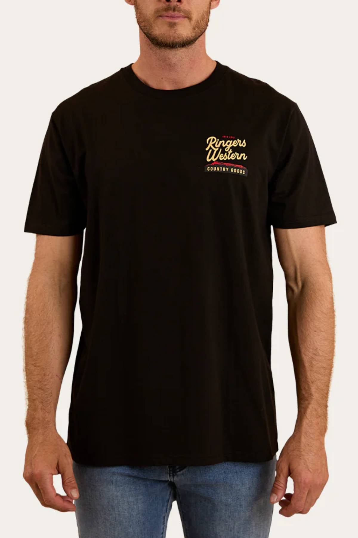 A man stands against a light background wearing the Ringers Western Country Goods Loose Fit T-Shirt, featuring yellow "Country Goods" text on the upper left chest, paired with blue jeans.
