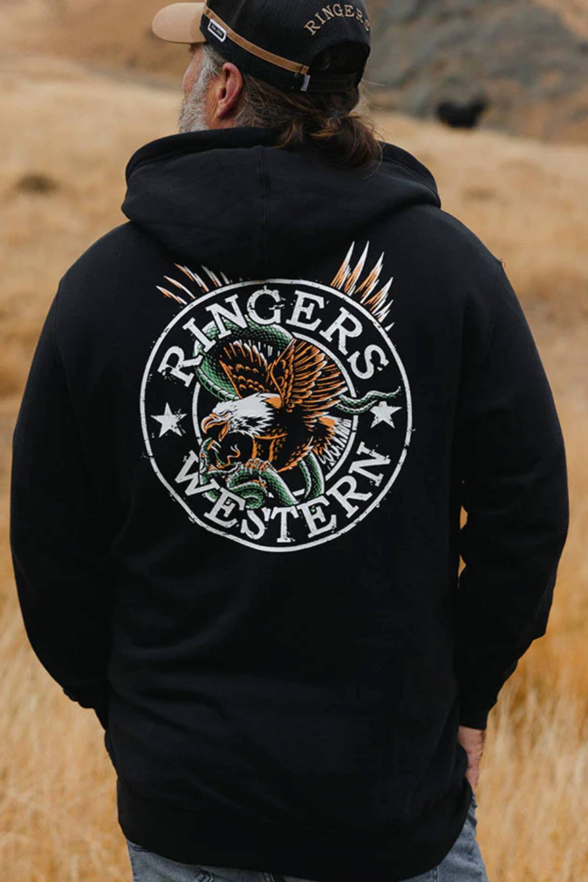 A man in a black Ringers Western Eyrie Pullover Hoodie and cap stands outdoors. The cozy brushed fleece hoodie displays a large eagle graphic and "Ringers Western" on the back amid a dry, grassy landscape.