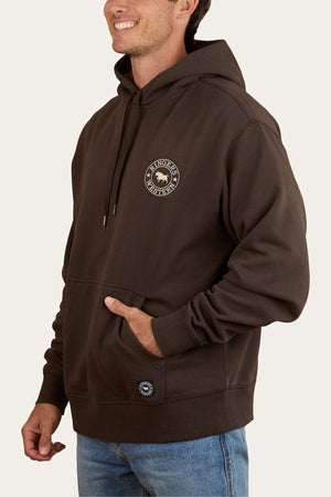 A man wearing a dark brown Ringers Western Get Your Truck On Pullover Hoodie, featuring premium brushed fleece, a small circular logo on the left chest, and a front pocket. He pairs it with blue jeans, smiles, and has one hand in the pocket.