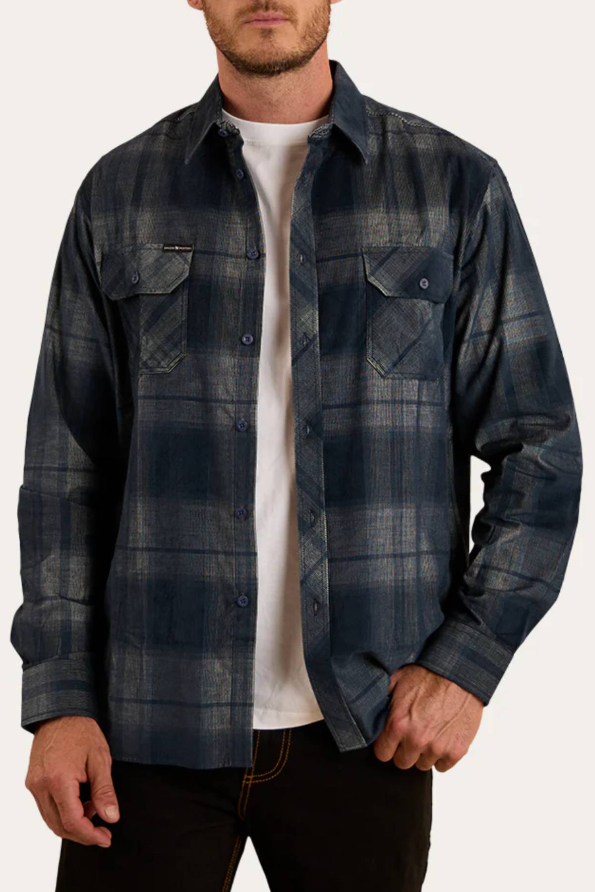 A man wears the Ringers Western Glenferrie Flannel Shirt in blue and gray plaid over a white t-shirt and black pants, one hand in his pocket. The 100% cotton shirt adds classic, laid-back style to his look.