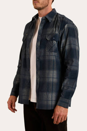 A man wears a Ringers Western Glenferrie Flannel Shirt in blue plaid, made from 100% cotton, over a white t-shirt with black pants, standing against a plain light background.