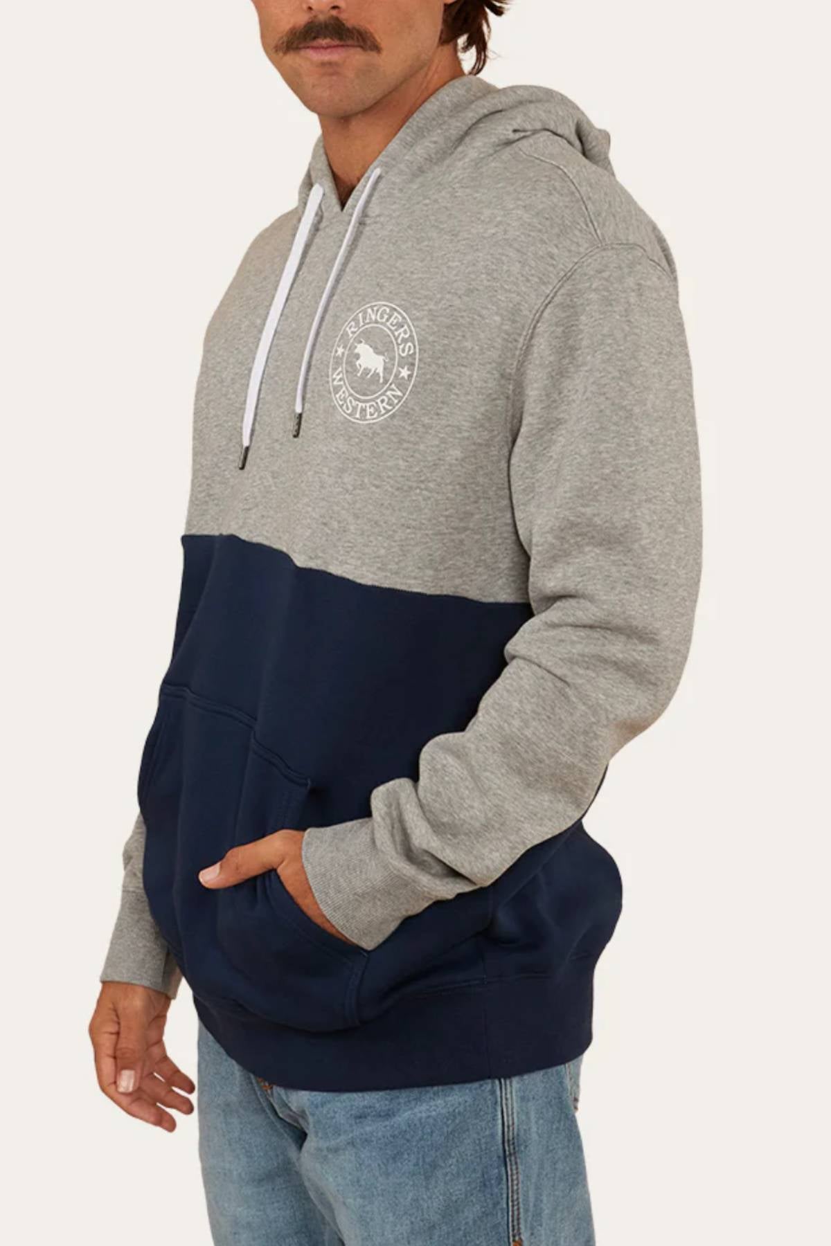 A man wears a Ringers Western Hahndorf Hoodie in a relaxed fit with a grey top, navy blue bottom, white chest emblem, white drawstring, and brushed fleece fabric. He pairs it with blue jeans and rests his left hand in the hoodie pocket.