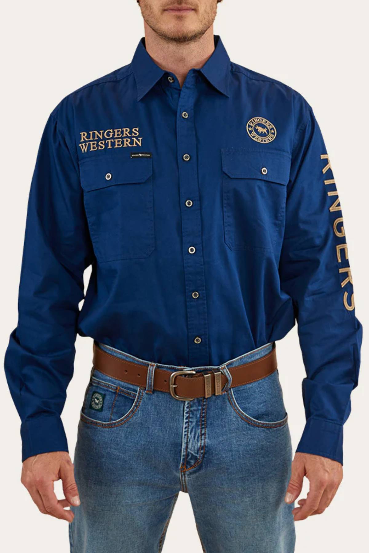 A man wears the Ringers Western Hawkeye Embroidered blue long-sleeve shirt with UPF50+ protection, "RINGERS WESTERN" text on the chest and sleeve, paired with blue jeans and a brown belt.