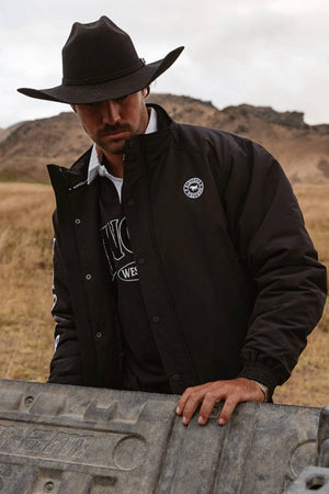 A man in a black cowboy hat and a Ringers Western Kidman Jacket stands in a grassy field with hills behind him, looking down and resting his hand on a metal surface.