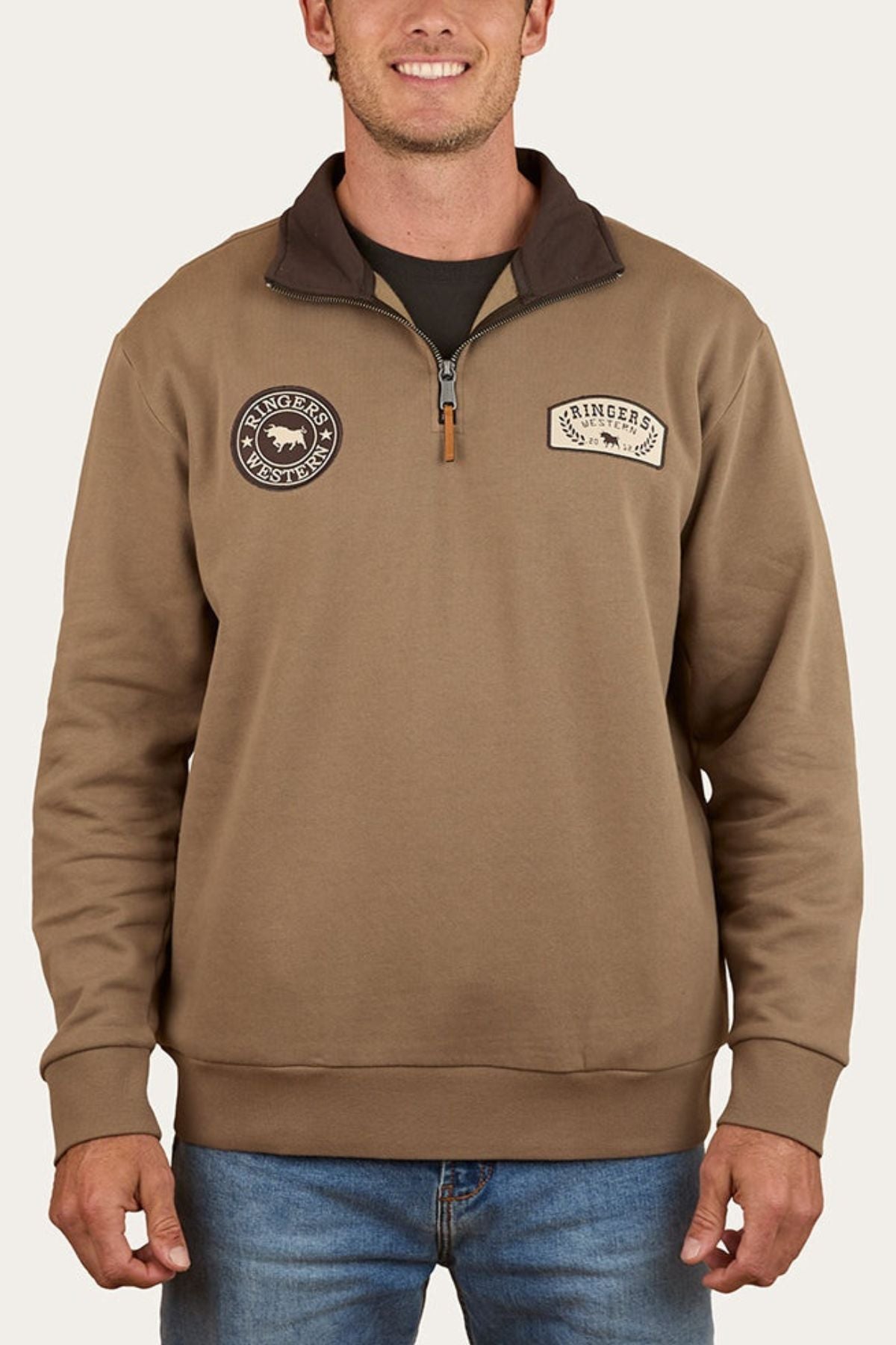 A smiling man wears the Ringers Western Lachlan 1/4 Zip Crew, a brown brushed fleece with two round chest patchesโone featuring a bull and brand textโpaired with blue jeans.