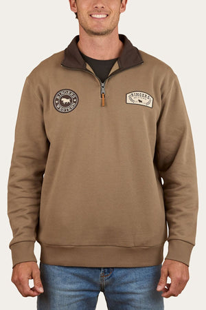 A smiling man wears the Ringers Western Lachlan 1/4 Zip Crew, a brown brushed fleece with two round chest patchesโone featuring a bull and brand textโpaired with blue jeans.