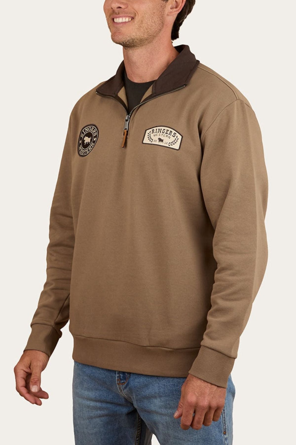 A man stands smiling in blue jeans and a Ringers Western Lachlan 1/4 Zip Crew with two chest patches.