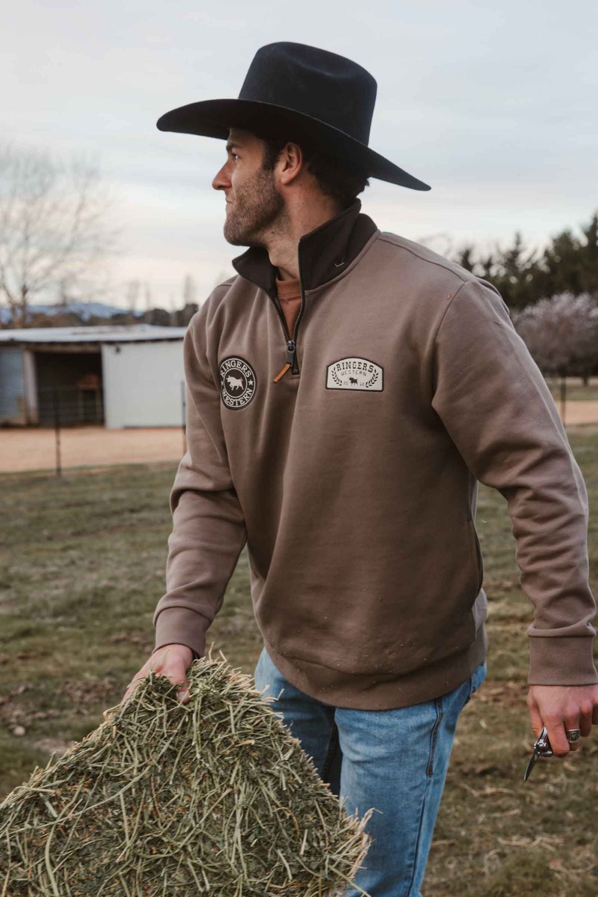 A man in jeans and a black cowboy hat wears a Ringers Western Lachlan 1/4 Zip Crew as he carries hay on a farm, with a barn and trees in the background.