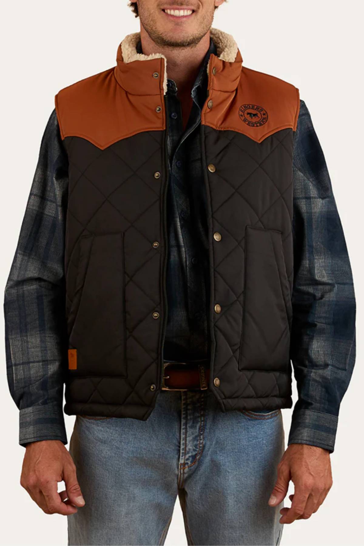 A man wears the Ringers Western McKinlay Puffer Vest, featuring brown shoulder panels, snap buttons, and a chest patch, over a blue plaid shirt and jeansβideal style for outdoor enthusiasts.