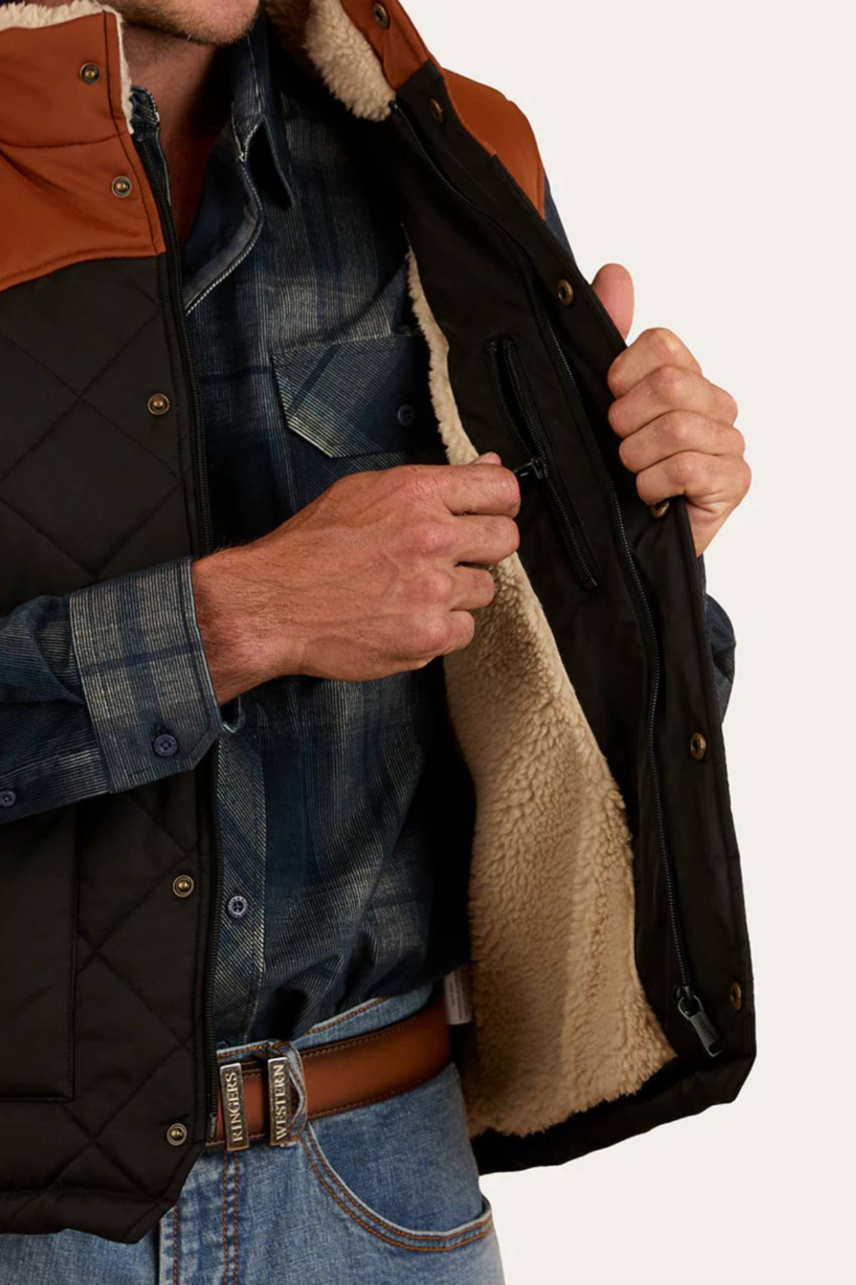 An outdoor enthusiast in a blue plaid shirt and jeans, wearing the Ringers Western McKinlay Puffer Vest with a brown belt, unzips an inner pocket to reveal its cozy sherpa lining.