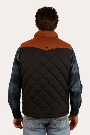 A man faces away, wearing the Ringers Western McKinlay Puffer Vest with a brown yoke over a blue plaid shirt and jeans. A small animal logo is displayed near the top center of the back, making it ideal for outdoor enthusiasts.