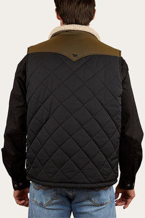 A person wears the Ringers Western McKinlay Puffer Vest with a black quilted design, olive green shoulder yoke, and subtle stitched pattern over a black long-sleeve shirt and blue jeans. The vest features the logo below the collar.