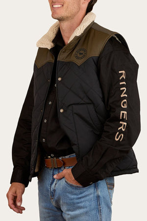 A man wears the Ringers Western McKinlay Puffer Vest—a black quilted vest with tan shoulders and a shearling collar, "RINGERS" on the sleeve. He smiles, left hand in his jeans pocket, facing slightly right.