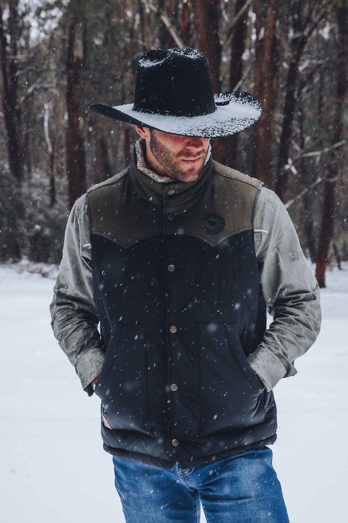 A man stands outdoors in falling snow, dressed in jeans, a winter jacket, a wide-brimmed black hat, and layered with the Ringers Western McKinlay Puffer Vest for extra warmth. Snow blankets the ground and trees as he keeps his hands in his pockets.