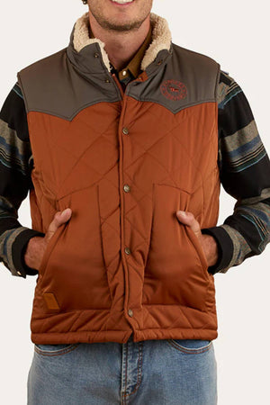 A person wearing the Ringers Western McKinlay Puffer Vest—brown with a gray yoke, shearling collar, and lightweight design—over a striped shirt, hands in vest pockets. Ideal for outdoor enthusiasts seeking comfort and style.