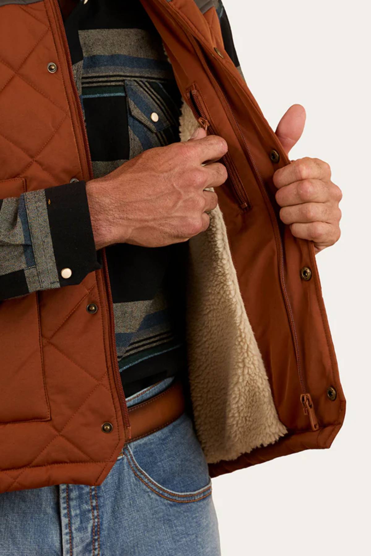 Wearing blue jeans and a striped shirt, a person shows off the Ringers Western McKinlay Puffer Vest—brown, quilted, with soft cream fleece lining, inner zip pocket, and lightweight design—ideal for outdoor enthusiasts.