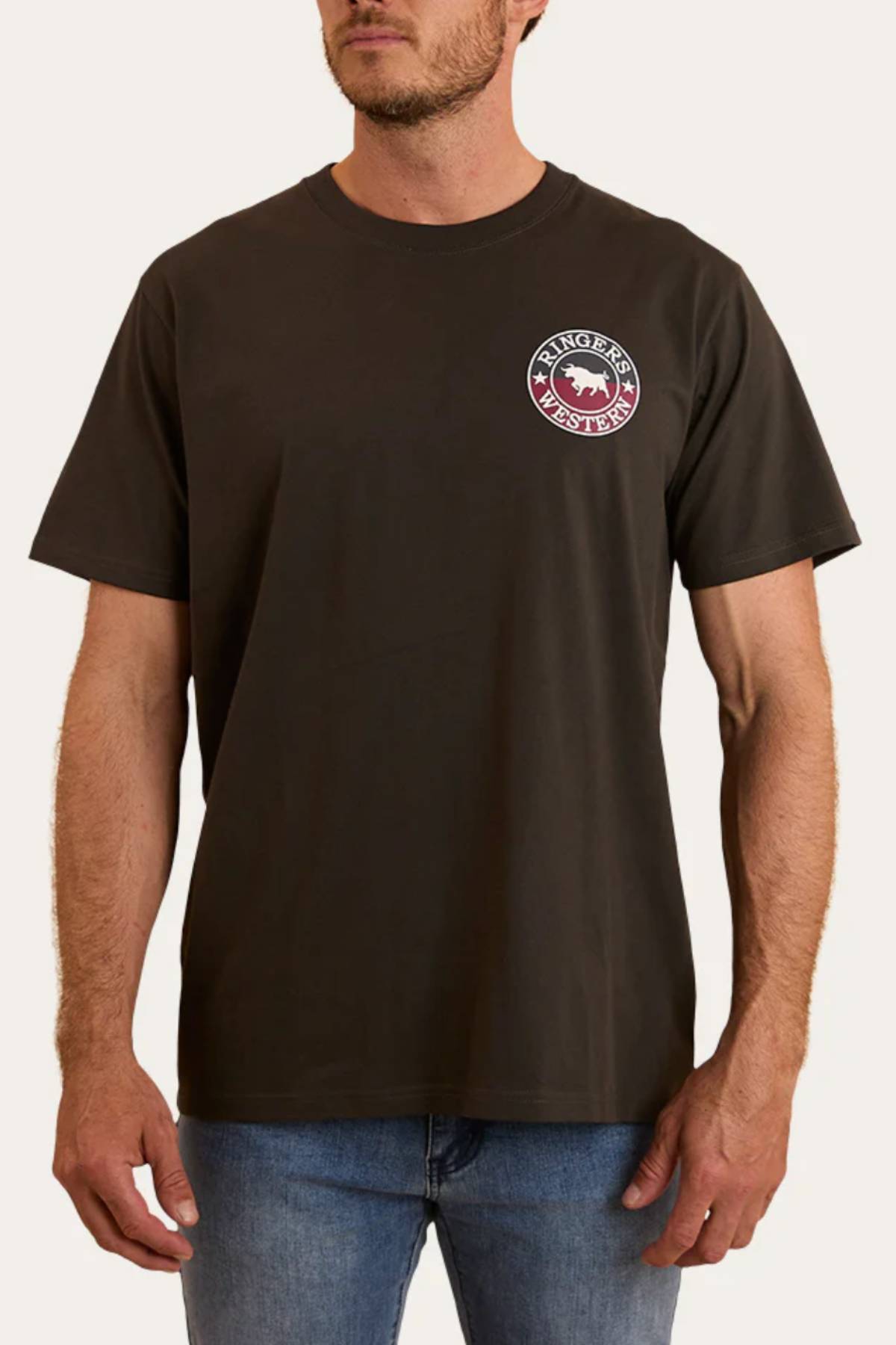 A man in a dark brown Ringers Western Signature Bull Loose Fit T-Shirt, made from 100% cotton jersey with a small circular chest graphic, and blue jeans stands against a plain light background.