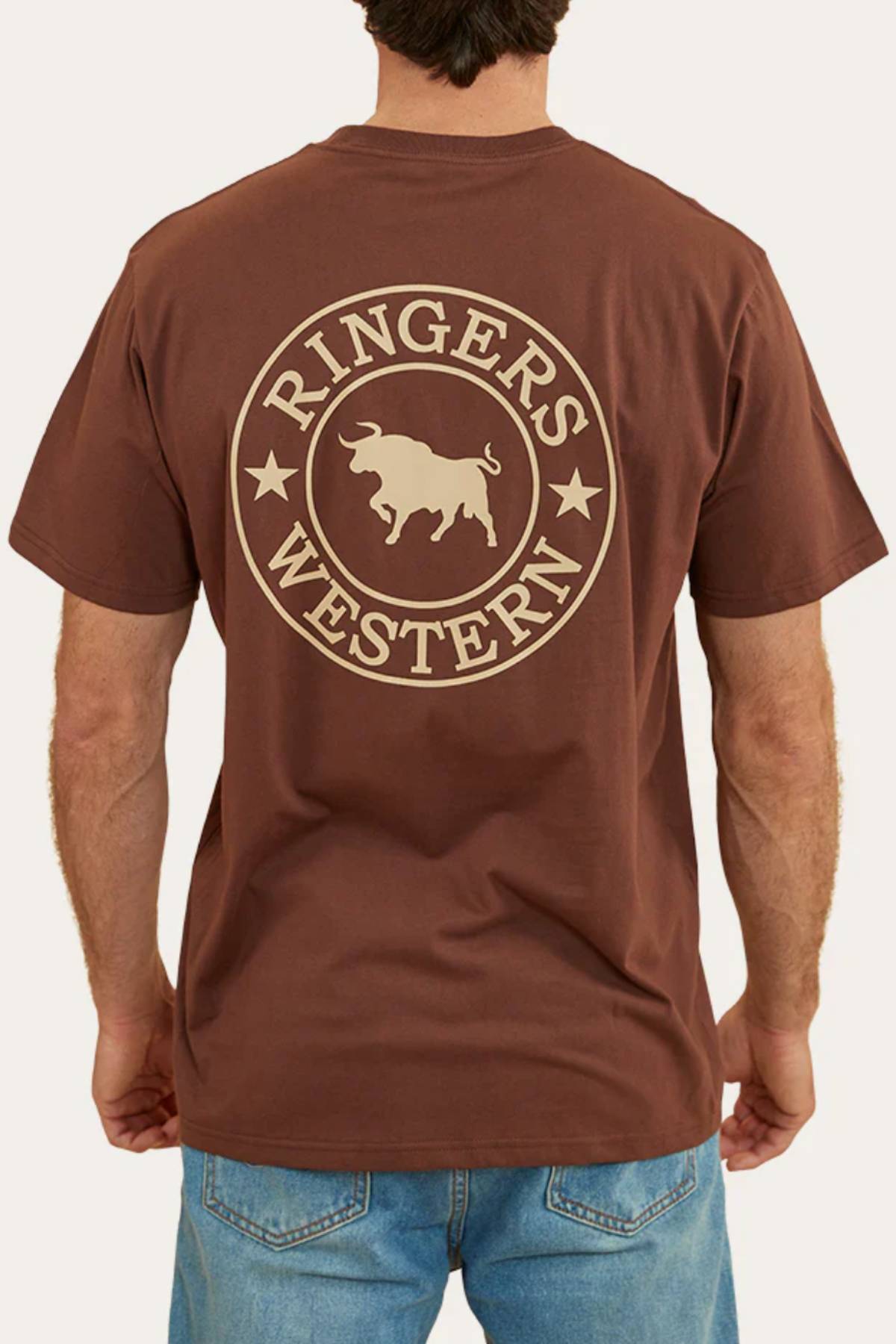 A person in blue jeans is seen from behind wearing a brown Ringers Western Signature Bull Loose Fit T-Shirt, made from 100% cotton jersey, with a large circular bull and two stars logo on the back.