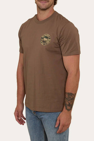 A man wears a brown Ringers Western Signature Bull Loose Fit T-Shirt (100% cotton jersey) with a small round logo on the left chest, paired with blue jeans. He has a tattoo on his left forearm and stands against a plain background.