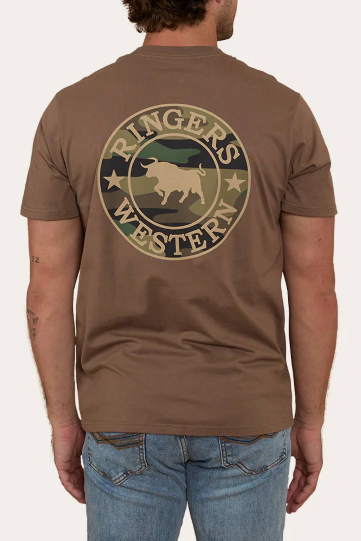 A man wears the Ringers Western Signature Bull Loose Fit T-Shirt in brown, featuring a circular bull and star logo on the back. Made from 100% cotton jersey, this tee pairs well with light blue jeans.