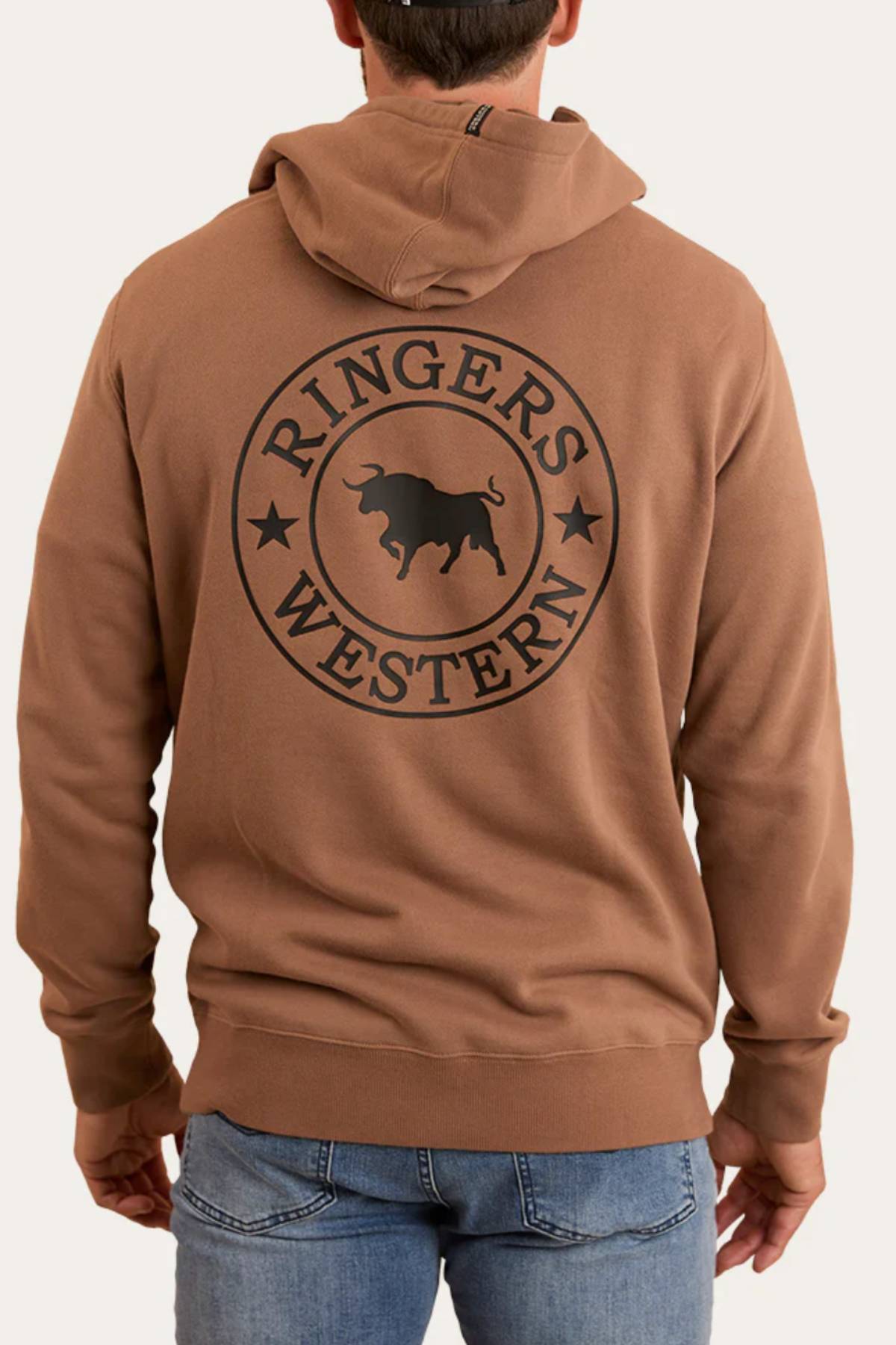 A man facing away from the camera wears blue jeans and a brown Ringers Western Signature Bull Pullover Hoodie featuring a large black logo and bull graphic on the back.