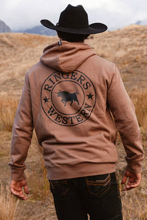A person stands outdoors facing away in a dry, grassy field, wearing a black cowboy hat and the Ringers Western Signature Bull Pullover Hoodie, featuring "Ringers Western" and a bull logo on the back.