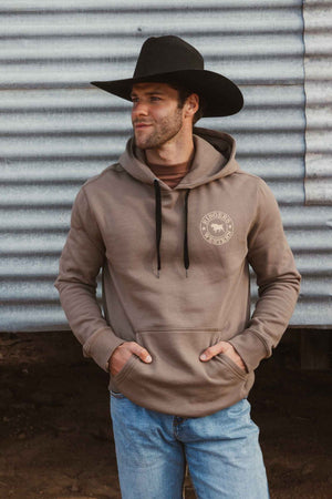 A man wearing a black cowboy hat, blue jeans, and a Ringers Western Signature Bull Pullover Hoodie with front pocket stands in front of a corrugated metal wall, looking to the side with his hands in his hoodie pocket.