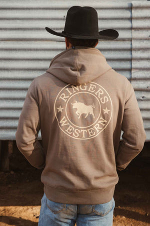 A person in a black cowboy hat and a brown Ringers Western Signature Bull Pullover Hoodie featuring a bull image on the back stands in front of a corrugated metal wall.